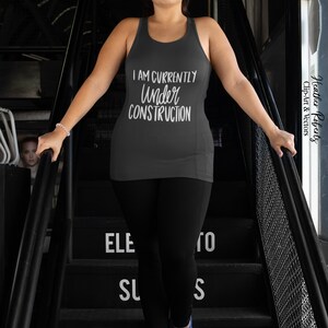 K&ouml;nnte beinhalten: Ein dunkelgraues Tanktop mit dem wei&szlig;en Schriftzug "I AM CURRENTLY UNDER CONSTRUCTION". Die Person tr&auml;gt schwarze Leggings und blaue und orangefarbene Turnschuhe. Das Bild ist auf einer schwarzen Treppe aufgenommen.