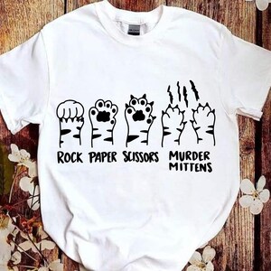 May include: White t-shirt with a graphic of cat paws in the shape of rock, paper, scissors, and murder mittens. The text "Rock Paper Scissors Murder Mittens" is printed below the paws.