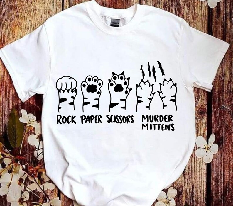 May include: White t-shirt with a graphic of cat paws in the shape of rock, paper, scissors, and murder mittens. The text "Rock Paper Scissors Murder Mittens" is printed below the paws.