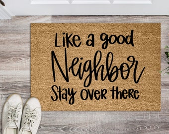 以下が含まれることがあります： 黒い文字で「Like a good neighbor stay over there」と書かれた茶色のココナッツ製のドアマット