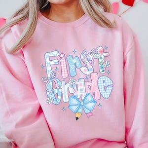 May include: A light pink sweatshirt with the words "First Grade" in a colorful, patchwork design. The letters are decorated with bows, flowers, and a pencil, perfect for back-to-school.