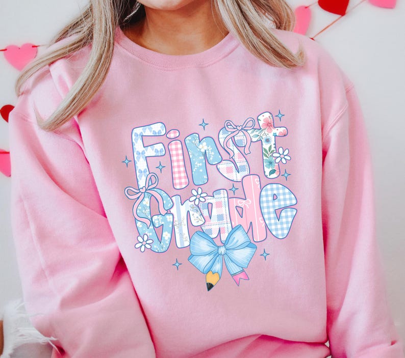 May include: A light pink sweatshirt with the words "First Grade" in a colorful, patchwork design. The letters are decorated with bows, flowers, and a pencil, perfect for back-to-school.