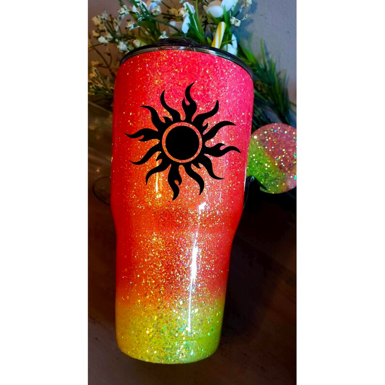 May include: A pink and yellow ombre glitter tumbler with a black sun design.
