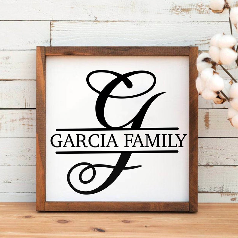 May include: A white wooden sign with a black cursive letter G and the text "Garcia Family" centered below the letter. The sign is framed in a dark brown wood frame.