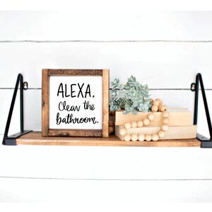 Puede incluir: Un letrero de madera con el texto "ALEXA, Limpia el ba&ntilde;o." sobre un fondo blanco. El letrero est&aacute; colocado en un estante de madera con una peque&ntilde;a planta y una cadena de cuentas de madera.