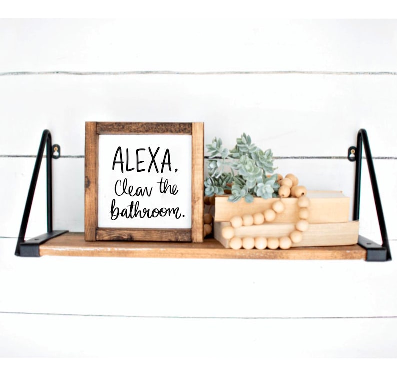 Puede incluir: Un letrero de madera con el texto "ALEXA, Limpia el ba&ntilde;o." sobre un fondo blanco. El letrero est&aacute; colocado en un estante de madera con una peque&ntilde;a planta y una cadena de cuentas de madera.
