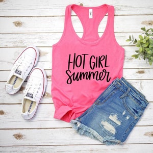 May include: A pink tank top with the text "HOT GIRL Summer" in black lettering. The tank top is laid out on a white wooden surface with a pair of white sneakers and blue denim shorts.