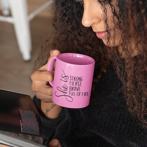 May include: A pink ceramic mug with black text that reads "She is strong fierce brave full of fire".