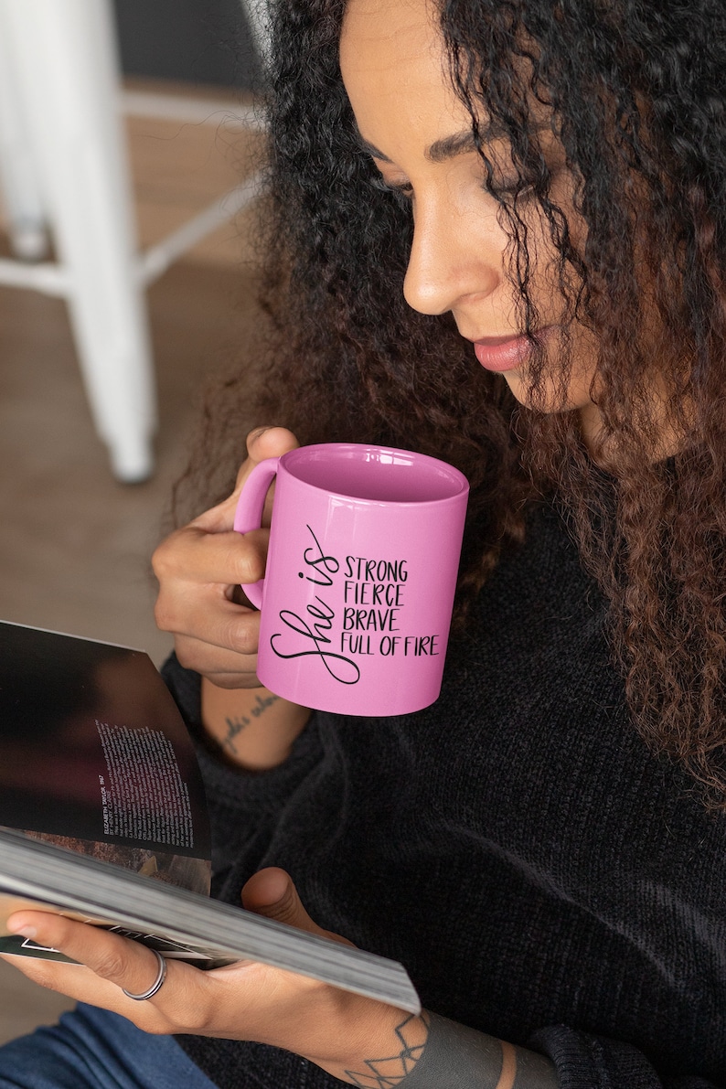 May include: A pink ceramic mug with black text that reads "She is strong fierce brave full of fire".