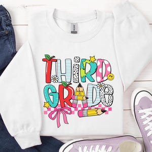 Puede incluir: Sudadera blanca con un gr&aacute;fico colorido que dice "Tercer Grado" con un arco&iacute;ris, l&aacute;pices y un patr&oacute;n a cuadros.