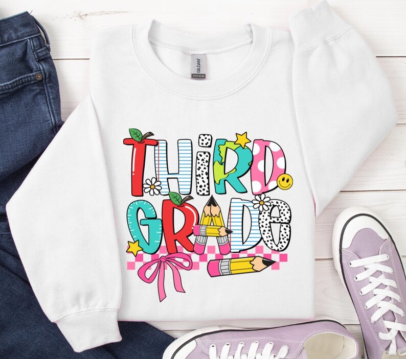 Puede incluir: Sudadera blanca con un gr&aacute;fico colorido que dice "Tercer Grado" con un arco&iacute;ris, l&aacute;pices y un patr&oacute;n a cuadros.