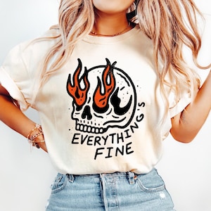 May include: Cream-colored t-shirt featuring a black skull with orange flames for eyes. The text "EVERYTHING'S FINE" is printed in black below the skull. The shirt has short sleeves and a crew neck.