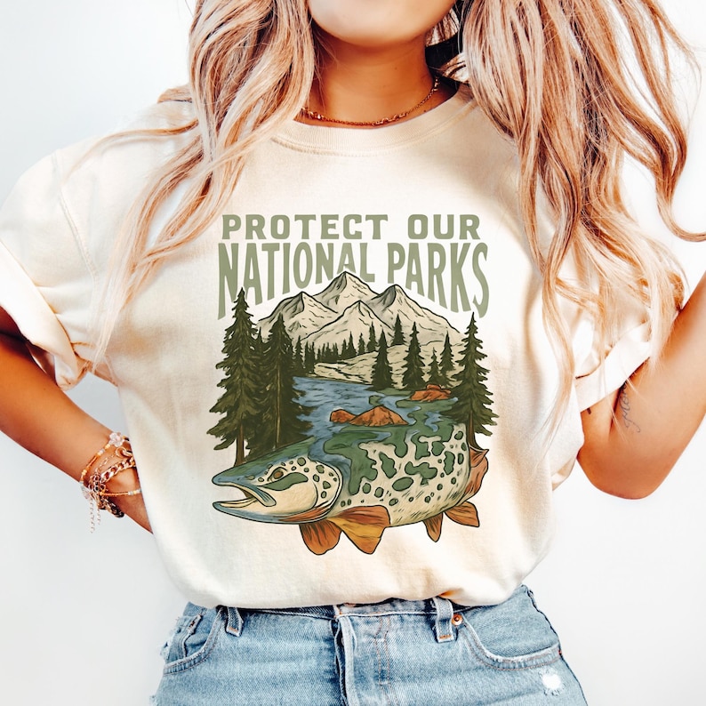 May include: Cream-colored sweatshirt with a graphic that reads "PROTECT OUR NATIONAL PARKS." The design features a mountain range, river, trees, and a fish. The sweatshirt is made of a soft material.