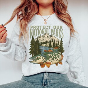 May include: A light gray sweatshirt featuring a graphic of a fish, river, mountains, and trees. The text "PROTECT OUR NATIONAL PARKS" is printed above the graphic. The sweatshirt is made of a soft material.