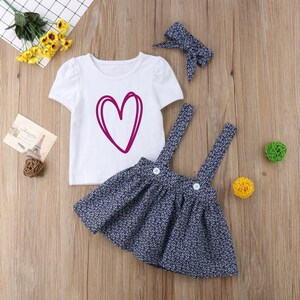 May include: A three-piece baby girl outfit with a white short-sleeved t-shirt featuring a pink heart design, a navy blue floral print overall skirt, and a pink bow headband.