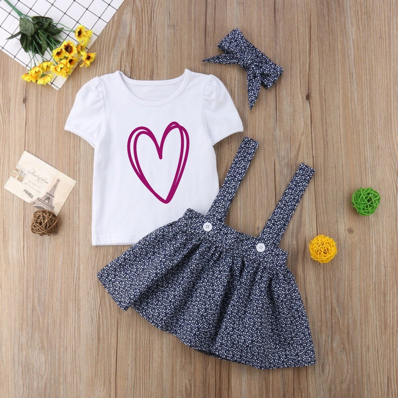 May include: A three-piece baby girl outfit with a white short-sleeved t-shirt featuring a pink heart design, a navy blue floral print overall skirt, and a pink bow headband.
