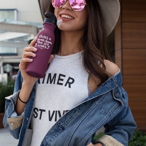 May include: A woman wearing a denim jacket, a white tank top with the word "SUMMER" printed on it, and a straw hat with a pink mirrored sunglasses. She is holding a purple water bottle with the text "There's a good chance this isn't water".