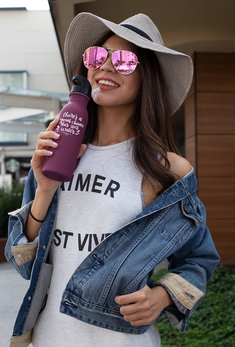 May include: A woman wearing a denim jacket, a white tank top with the word "SUMMER" printed on it, and a straw hat with a pink mirrored sunglasses. She is holding a purple water bottle with the text "There's a good chance this isn't water".