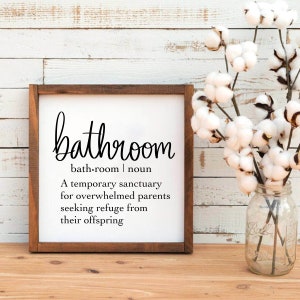 Puede incluir: Un letrero blanco con un marco de madera marr&oacute;n, que presenta la palabra "bathroom" en una fuente decorativa, con la definici&oacute;n "bath room | noun A temporary sanctuary for overwhelmed parents seeking refuge from their offspring".
