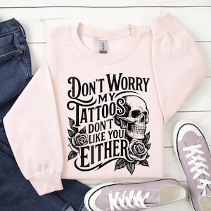 May include: A light pink sweatshirt with the text "Don't Worry My Tattoos Don't Like You Either" in black, along with a skull and roses graphic. The sweatshirt is paired with blue jeans and light purple sneakers.