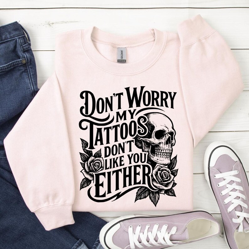 May include: A light pink sweatshirt with the text "Don't Worry My Tattoos Don't Like You Either" in black, along with a skull and roses graphic. The sweatshirt is paired with blue jeans and light purple sneakers.