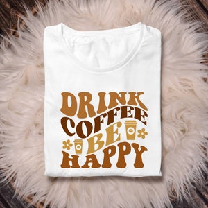 May include: White t-shirt with brown text that reads "Drink Coffee Be Happy" in a retro font. The text is surrounded by brown coffee cup illustrations.
