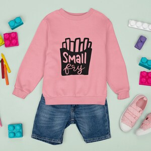 May include: Pink sweatshirt with a black graphic of french fries and the text "Small fry". The sweatshirt is paired with blue denim shorts and pink polka dot sneakers.
