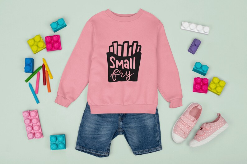 May include: Pink sweatshirt with a black graphic of french fries and the text "Small fry". The sweatshirt is paired with blue denim shorts and pink polka dot sneakers.