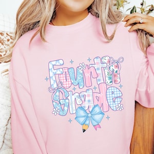 May include: A light pink sweatshirt with the words "Fourth Grade" in a colorful, patterned design. The letters are decorated with gingham, floral, and bow accents, with a blue pencil and bow at the bottom. The sweatshirt is a comfortable, casual style.
