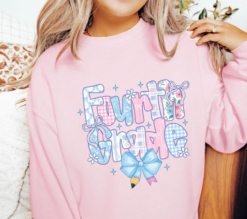 May include: A light pink sweatshirt with the words "Fourth Grade" in a colorful, patterned design. The letters are decorated with gingham, floral, and bow accents, with a blue pencil and bow at the bottom. The sweatshirt is a comfortable, casual style.