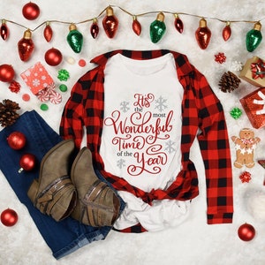 May include: A white t-shirt with red text that says "It's the most Wonderful Time of the Year". The shirt is lying on a white surface with a red and black checked shirt, blue jeans, and Christmas decorations.