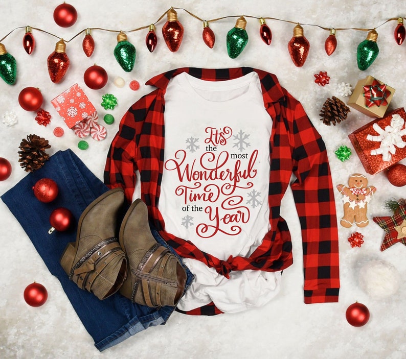May include: A white t-shirt with red text that says "It's the most Wonderful Time of the Year". The shirt is lying on a white surface with a red and black checked shirt, blue jeans, and Christmas decorations.