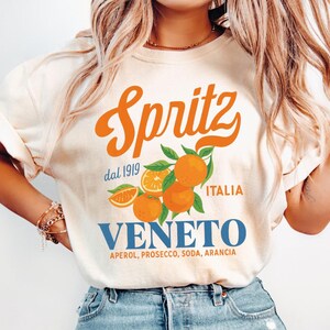 Könnte beinhalten: Cremefarbenes T-Shirt mit dem Wort "Spritz" in orangefarbener Schrift, mit Orangenillustrationen und dem Text "Veneto Italia". Das Shirt enthält auch den Text "dal 1919" und eine Zutatenliste.