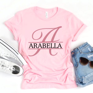May include: Pink t-shirt with a large, cursive letter "A" in a dusty rose color. The letter is split by a black horizontal line with the name "ARABELLA" printed in black letters.