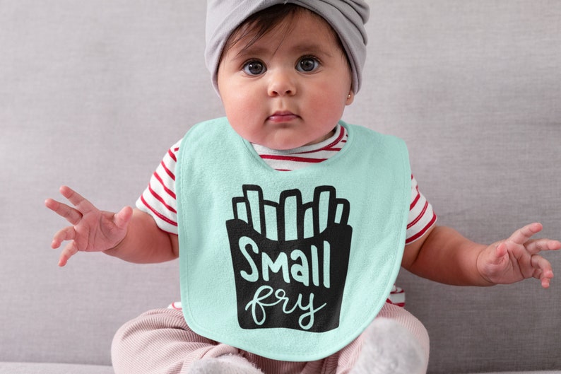 May include: A baby wearing a light blue bib with a black graphic of french fries and the text "Small fry". The baby is wearing a red and white striped shirt and a gray hat.