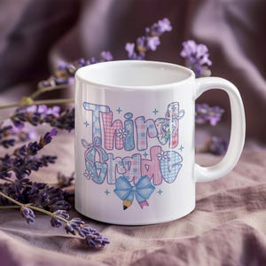 Puede incluir: Taza de cer&aacute;mica blanca con la frase "Third Grade" en un dise&ntilde;o colorido y estampado. Las letras est&aacute;n decoradas con motivos de cuadros, flores y lazos. Un lazo azul y un l&aacute;piz gr&aacute;fico en la parte inferior.