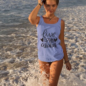 May include: A woman wearing a light blue tank top with the text "live love aloha" and two black hearts on the front. She is standing in the ocean with waves splashing around her.
