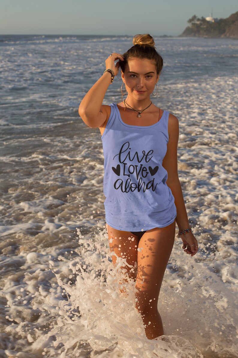 May include: A woman wearing a light blue tank top with the text "live love aloha" and two black hearts on the front. She is standing in the ocean with waves splashing around her.