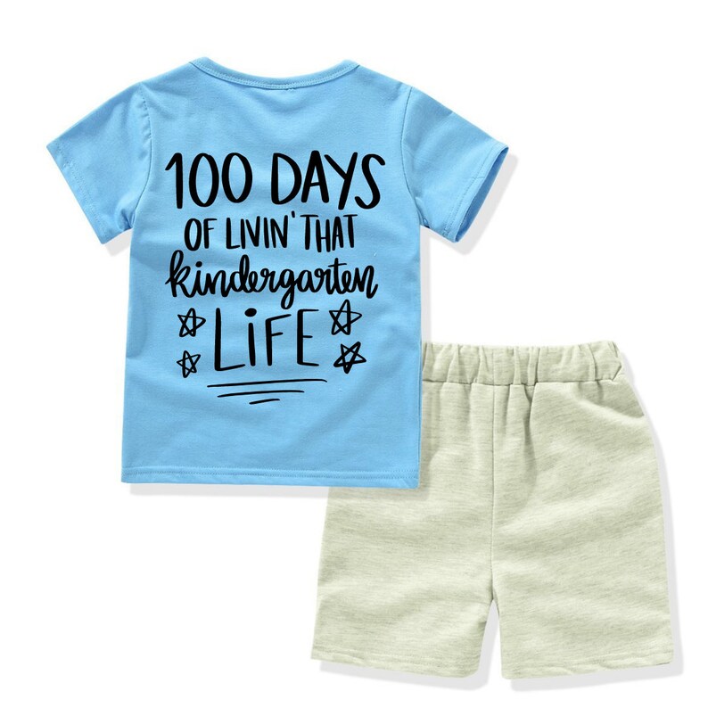 May include: A light blue short-sleeved t-shirt with the text "100 Days of Livin' That Kindergarten Life" in black. The shirt is paired with light gray shorts.