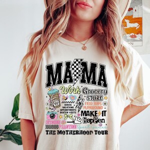 May include: A white t-shirt with a graphic design that says "Mama Work Grocery Store Field Trips Playground Make It Happen The Motherhood Tour". The graphic design is in black, pink, blue, and yellow.