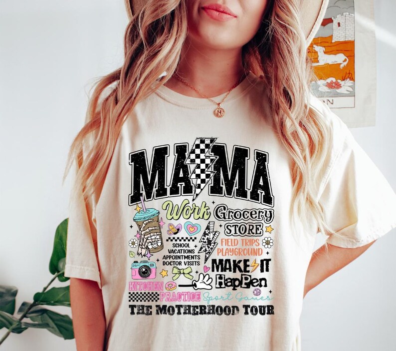May include: A white t-shirt with a graphic design that says "Mama Work Grocery Store Field Trips Playground Make It Happen The Motherhood Tour". The graphic design is in black, pink, blue, and yellow.