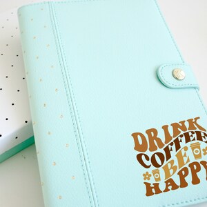 May include: A light blue leather planner with gold polka dots and a brown vinyl sticker that says "Drink Coffee Be Happy" with coffee cup illustrations.