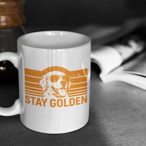 May include: White ceramic mug with an orange graphic of a Golden Retriever wearing sunglasses, set against a retro striped background. The text "STAY GOLDEN" is printed below the dog. A coffee carafe and open book are in the background.