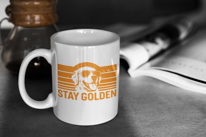 May include: White ceramic mug with an orange graphic of a Golden Retriever wearing sunglasses, set against a retro striped background. The text "STAY GOLDEN" is printed below the dog. A coffee carafe and open book are in the background.