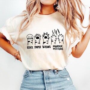 May include: White t-shirt with a graphic of cat paws in the shape of rock, paper, scissors, and murder mittens. The text "Rock Paper Scissors Murder Mittens" is printed below the graphic.