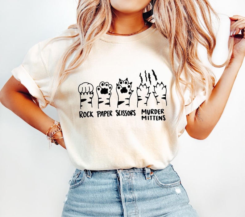 May include: White t-shirt with a graphic of cat paws in the shape of rock, paper, scissors, and murder mittens. The text "Rock Paper Scissors Murder Mittens" is printed below the graphic.