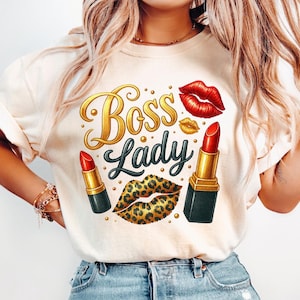 May include: A cream-coloured t-shirt with the words "Boss Lady" in gold and green script, featuring red lipstick, leopard print lips, and gold lips. The shirt is made of a soft material.