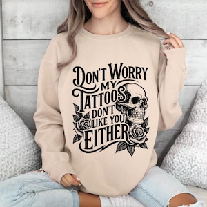 May include: Beige sweatshirt with the text "Don't Worry My Tattoos Don't Like You Either" in black, along with a skull and rose graphic. The sweatshirt is a neutral color and features a crew neck.