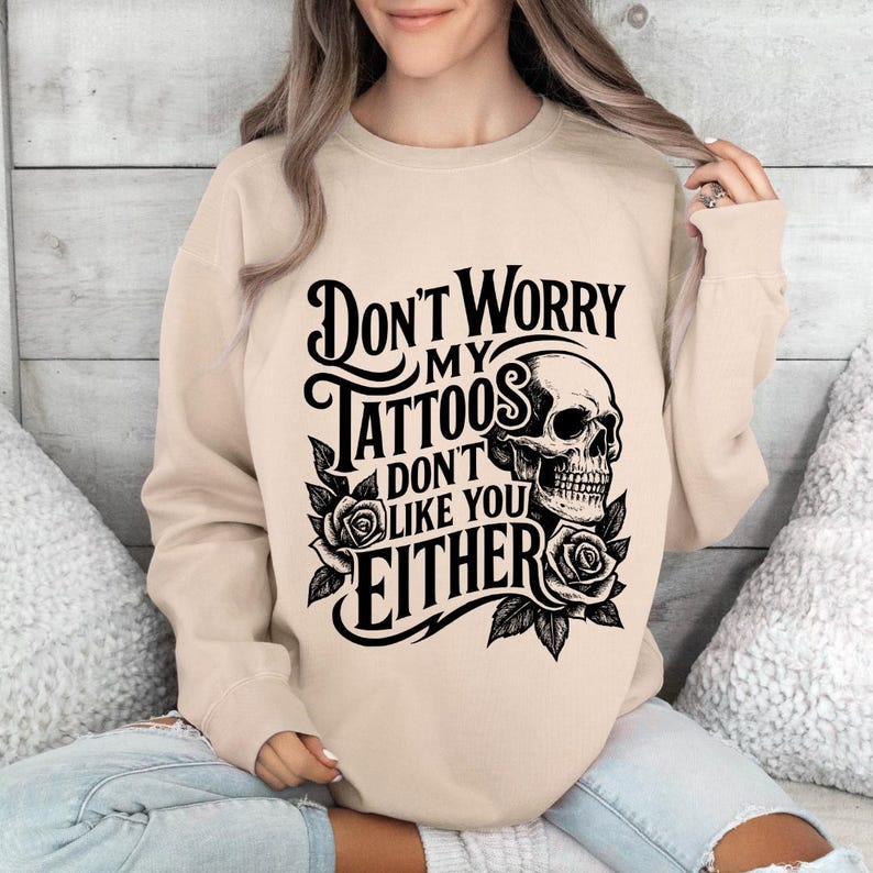 May include: Beige sweatshirt with the text "Don't Worry My Tattoos Don't Like You Either" in black, along with a skull and rose graphic. The sweatshirt is a neutral color and features a crew neck.