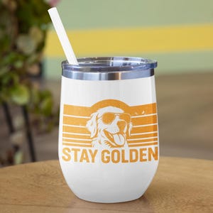 May include: White insulated tumbler with a silver rim and a white straw. The tumbler features an orange graphic of a Golden Retriever wearing sunglasses, with the words "STAY GOLDEN" below the dog.